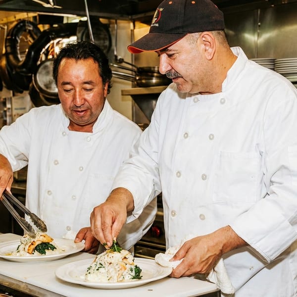Two chefs in white coats plating dishes with tongs in a bustling professional kitchen.